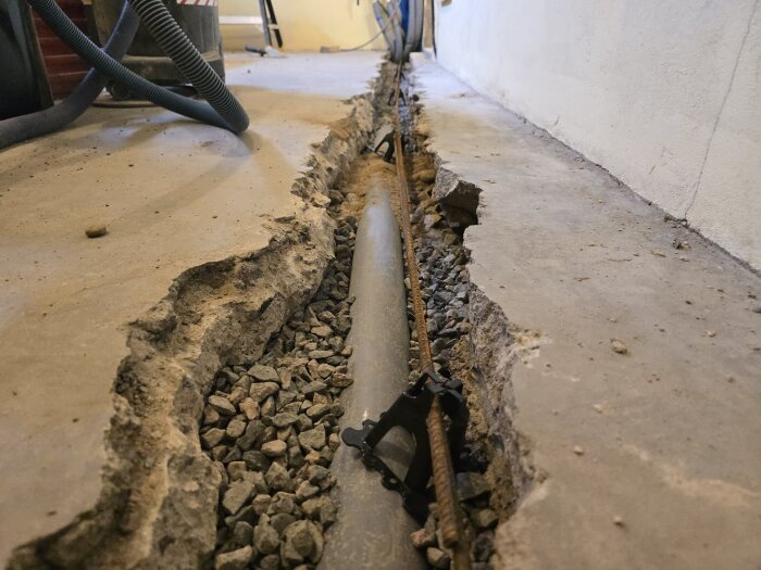 Ränna i golv med dränering och armeringsjärn, omgiven av grus och grov kant, i ett hobbyrum under renovering.
