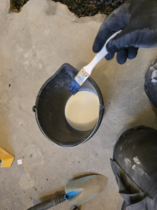 Person målar betonginsats med primer i en svart hink och håller en pensel med blå borst. Golvet är grått och det finns en spade närvarande.