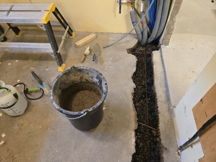 Hink med betong bredvid skåra i golvet, omgiven av verktyg och en pall, förberett för gjutning av avloppsränna i hobbyrum.