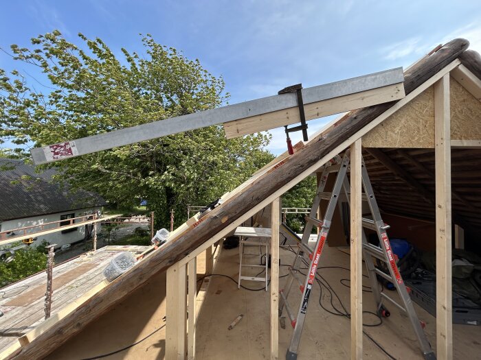 Takomläggning pågår med kupa mellan takstolar. En stege står lutad mot konstruktionen. Klar väderdag syns i bakgrunden.