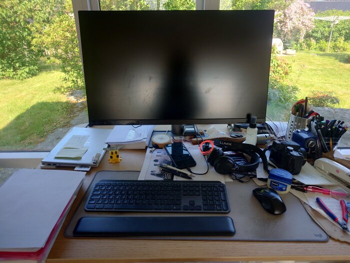 Arbetsbord med dator, skärm, tangentbord, hörlurar och skrivmaterial, beläget framför ett fönster med utsikt över en gräsmatta och buskar.