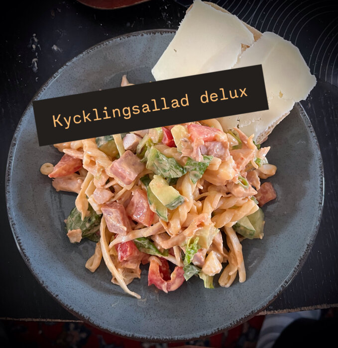 Kycklingsallad med grönsaker, pasta, ägg, landjäger, dressing och två skivor nybakat bröd med ost på en tallrik.