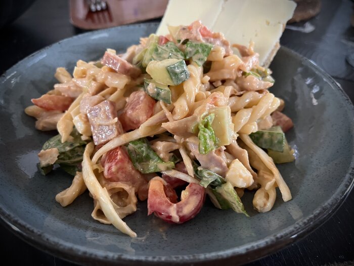 Kycklingsallad med pasta, grönsaker, landjäger och dressing på en tallrik, serverad med två skivor bröd i bakgrunden.