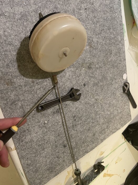 Reservdel av metall med handtag och skruvar, något som kan vara del av en reparation, ligger på en grå yta.