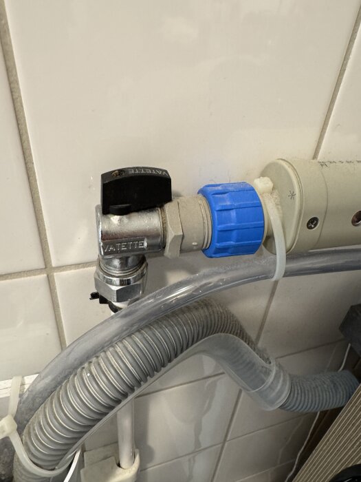 Kulventil med svart handtag och blå anslutning, kopplad till en 12 mm kopparrörsinstallation för tvättmaskin, mot vit kakelvägg.