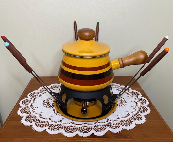 Gul fonduegryta med fyra fonduegafflar på en bord med spetsduk.