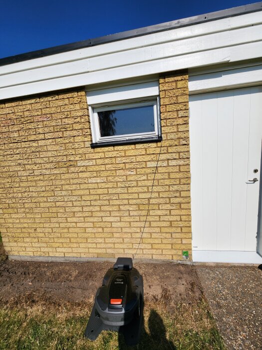 Gul tegelvägg med ett fönster och en vit dörr. En robotgräsklippare står på marken med ett draget kabelspår framför.