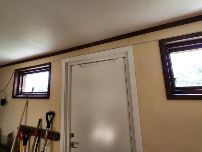 Vägg med två små fönster och en vit dörr, samt verktyg lutade mot väggen i ett garage.