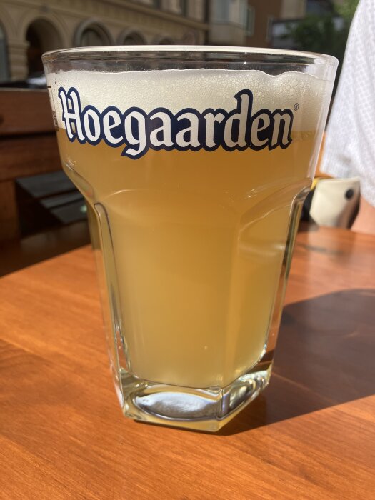 Ölglas med skummande Hoegaarden står på ett träbord i solen, bakgrund med suddiga byggnader.
