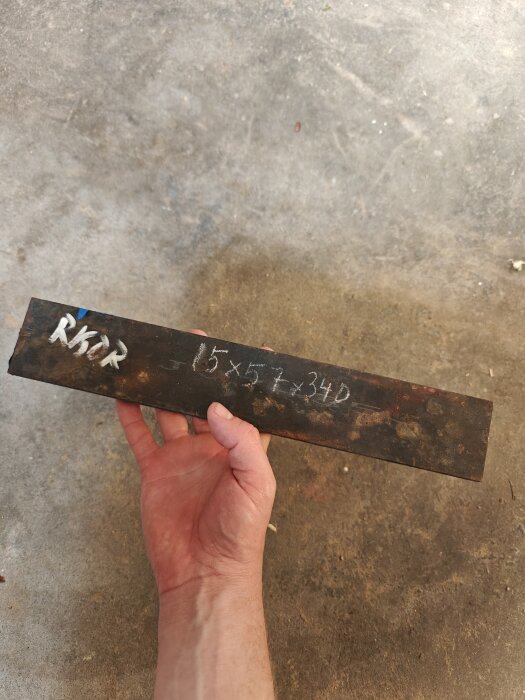 Hand håller en rostig metallbit märkt "Rigor" med måttangivelser.