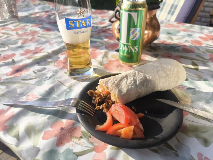Tallrik med taco-wrap och tomatklyftor, ölglas bredvid på blommigt dukat bord i solsken.