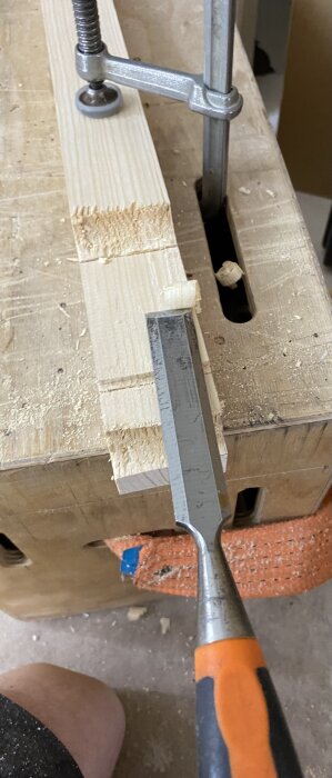 Trälist fäst med tving, bearbetad med stämjärn på en arbetsbänk. Verktygskista i bakgrunden.