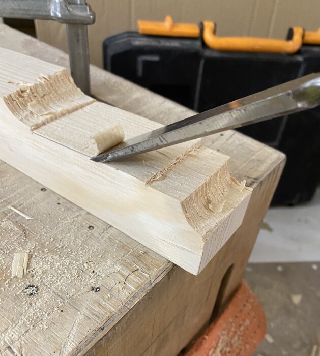 Träbit hålls i skruvstycke, bearbetad med stämjärn. Bakgrund: svart verktygslåda med orange handtag. Flisor och träspån på bänken.