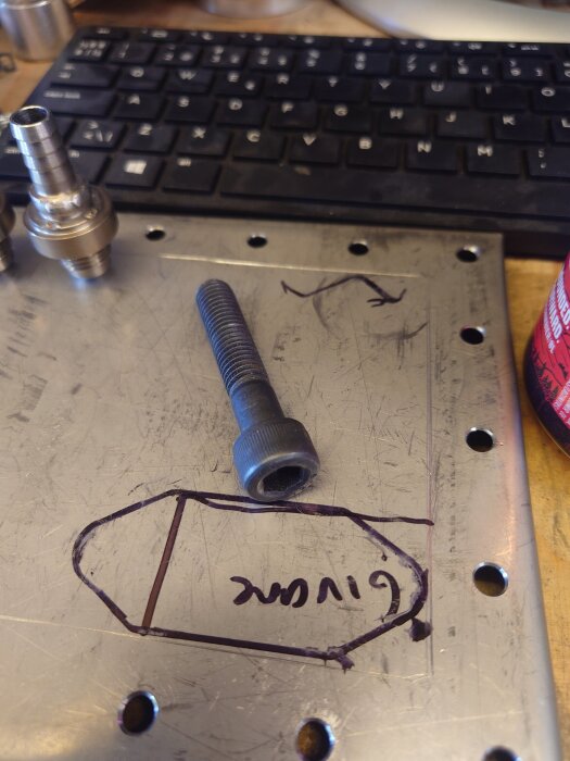 Skruv av klass 12.9 ligger på metallplåt vid tangentbord, med ritning och text "givare" på plåten.
