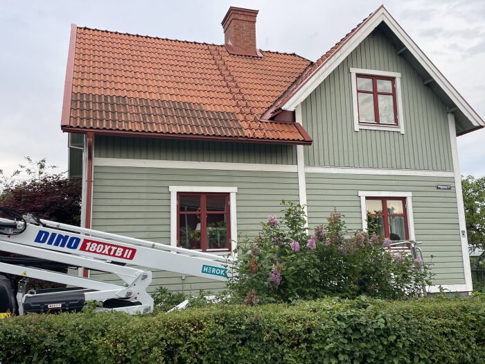 Grönt trähus med orange lertegeltak, synlig skorstensdel. Försök till rengöring av taket med högtryckstvätt visas. Vissa tegelytor är mörkare.