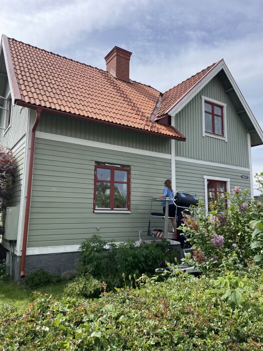 Grönt trähus med nylagt tegeltak, norrsidan behöver rengöras. Person på stege arbetar med att tvätta ytan med högtryckstvätt.