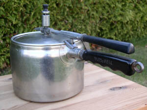 Tryckkokare från 60-talet med två svarta handtag, stående på ett träbord utomhus. Används för snabb tillagning av viltkött och potatis.