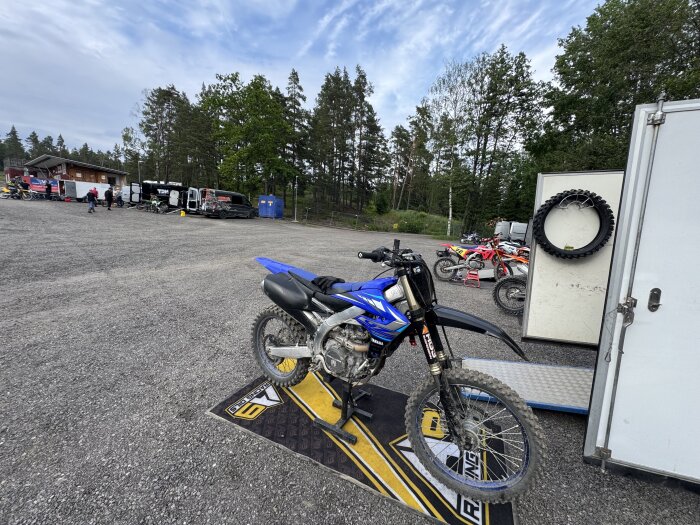 Blå motorcykel parkerad på en grusplan vid en motocrossbana, omgiven av släpvagnar och fler motorcyklar.