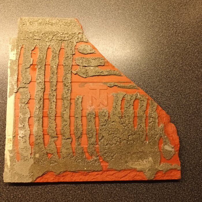 Ett hörn av ett rött klinker med ett inpräglat märke "TN" synligt på baksidan, kantat av rester av lim eller fogmassa.