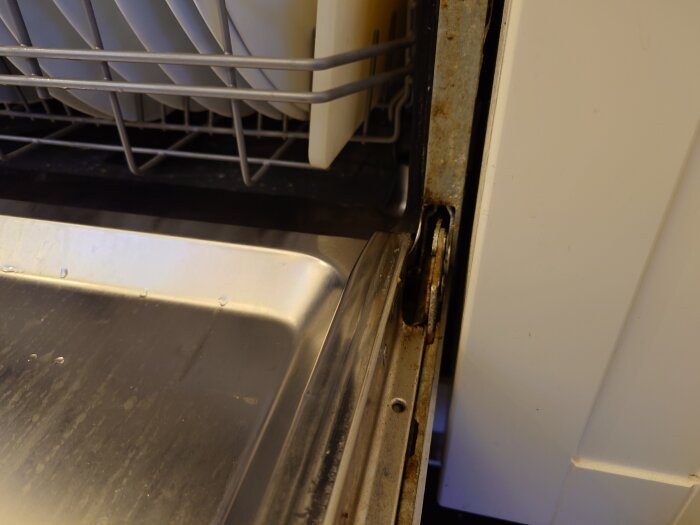 Närbild av en diskmaskinslucka med synligt gångjärn; insidan visar diskmaskinkorg med blött metallgolv.