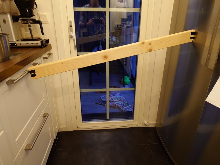 En träregel spänner över en nerfälld diskmaskin i ett kök, för att hålla luckan stängd. Kaffebryggare och kylskåp syns i bakgrunden.