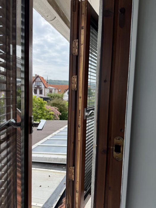 Öppnat träfönster med monterad persienn, utsikt över tak och trädgård.