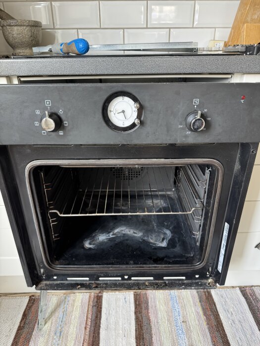 Ugn med öppnad lucka, två vred och en klocka på framsidan, blå verktyg ovanpå, köksmiljö med vita kakelplattor i bakgrunden.