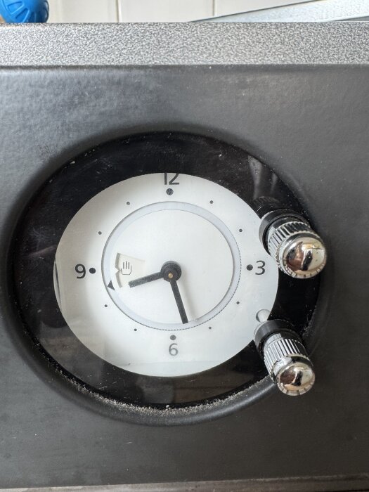 Klocka på ugnspanel med två rattar och en urtavla som visar en hand-ikon, relaterad till spärrefunktion för ugnsstart.