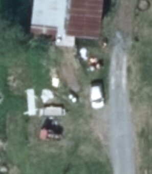 Markområde med grusväg, grönska, en byggnad med plåttak och en vit bil parkerad bredvid material och föremål på marken.