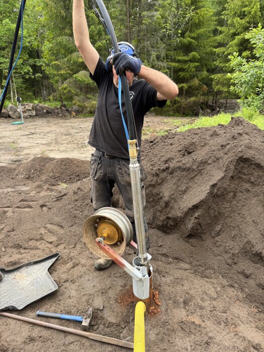 Person som drar upp en gammal pump ur marken i skogsområde, omgiven av jord och verktyg.