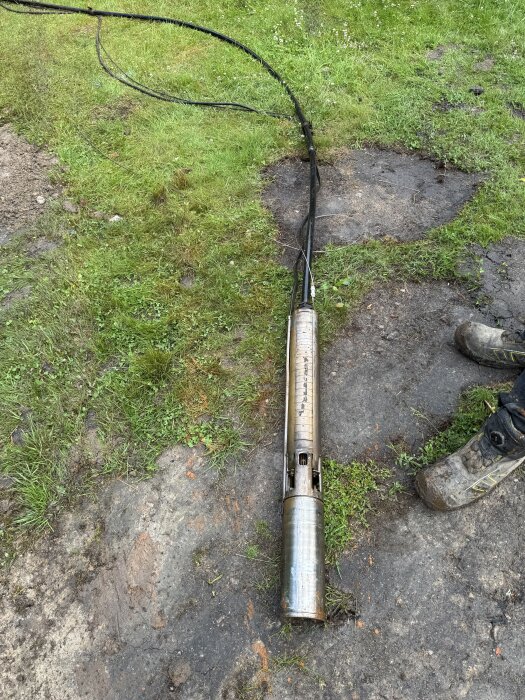 En gammal vattenpump ligger på marken bredvid kablar, omgiven av gräs och jordig yta. En stövel syns delvis i bild.