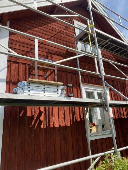 Röd trävägg med ställningar och vita foder runt ett fönster, soligt väder.