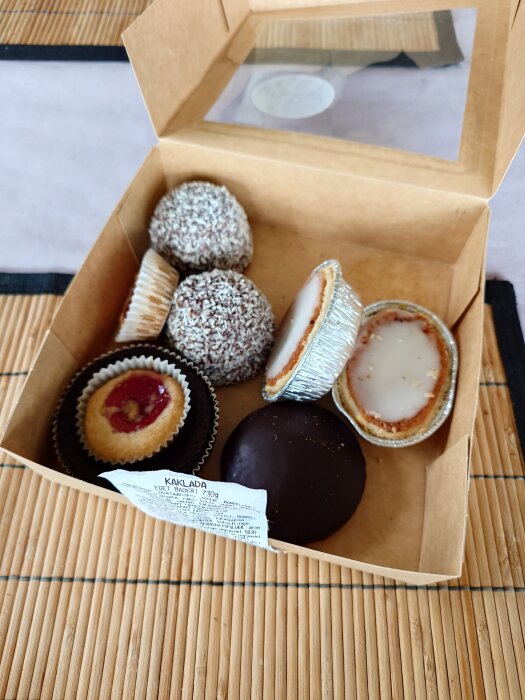 En öppen kartong med olika bakverk, inklusive chokladbollar, mandelformar med glasyr, och kakor med sylt på en bordstablett av bambu.