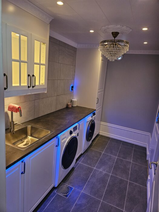 Tvättstuga med inbyggda tvättmaskiner under en bänkskiva, kaklade väggar och svart klinkergolv. En diskbänk och elegant taklampa syns också.