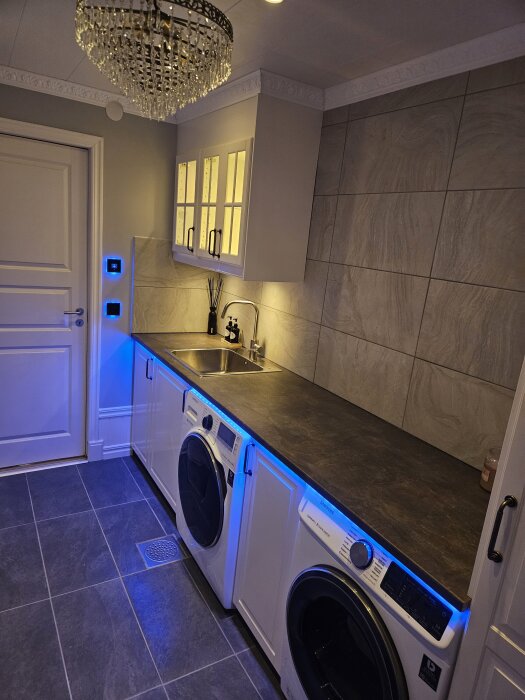 Tvättstuga med inbyggda tvättmaskiner under en stor bänkskiva, kaklade väggar, kristallkrona och belysning.