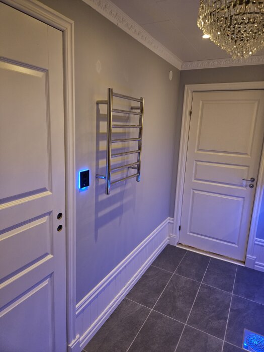 Grå tvättstuga med vita dörrar, stålrör till handdukstork, mörkgrått klinkergolv och glittrig lampa i taket.