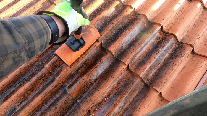 En person rengör ett smutsigt tak av terrakottafärgade tegelpannor med en handske och en borste.