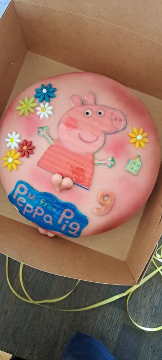 Rosa tårta med en illustration av en gris, omgiven av färgglada blommor och texten "Peppa Pig" samt siffran 9.