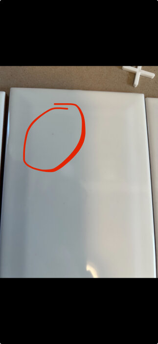 Vit kakelplatta med små orangea prickar under glasyren, markerad med en röd cirkel.