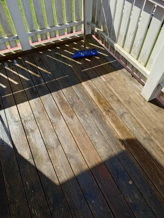 En smutsig balkong med trätrall i svart-grå-brun nyans och en blå borste, före rengöring. Skugga från räcke syns på golvet.