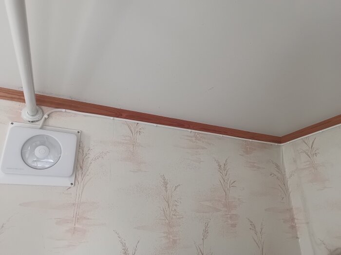 Vägg med blommig tapet och trälist upptill. En vit ventilationsfläkt monterad på väggen nära taket.
