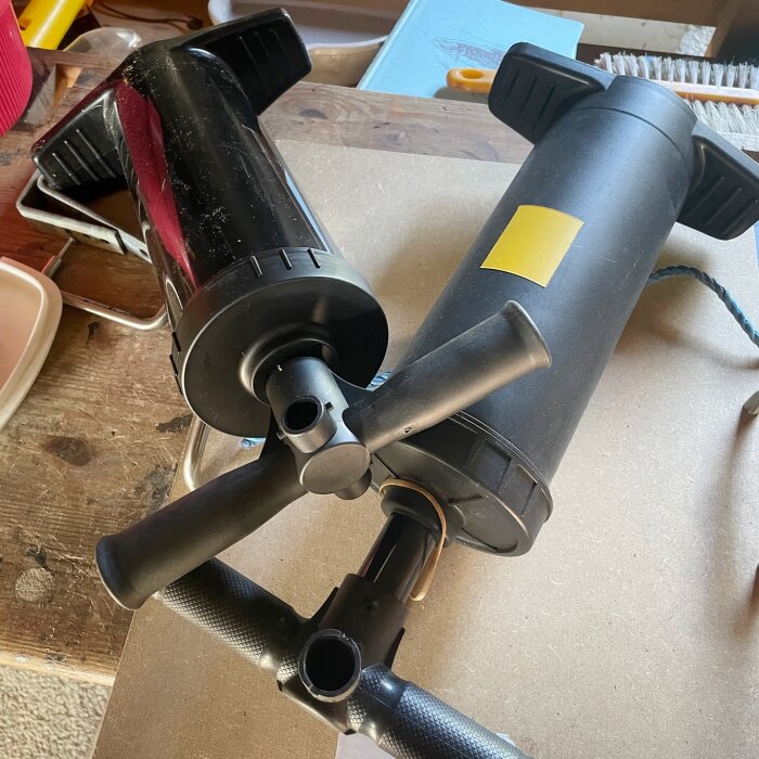 Två svarta dubbelverkande handpumpar på en arbetsbänk, en med gul etikett, tillsammans med olika verktyg och material i bakgrunden.