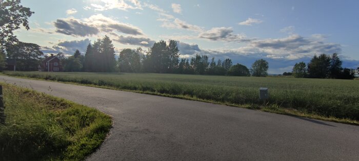 Landskapsvy med asfalterad väg, fält och ett rött trähus omringat av träd under en molnig himmel. Solstrålar skiner genom molnen.