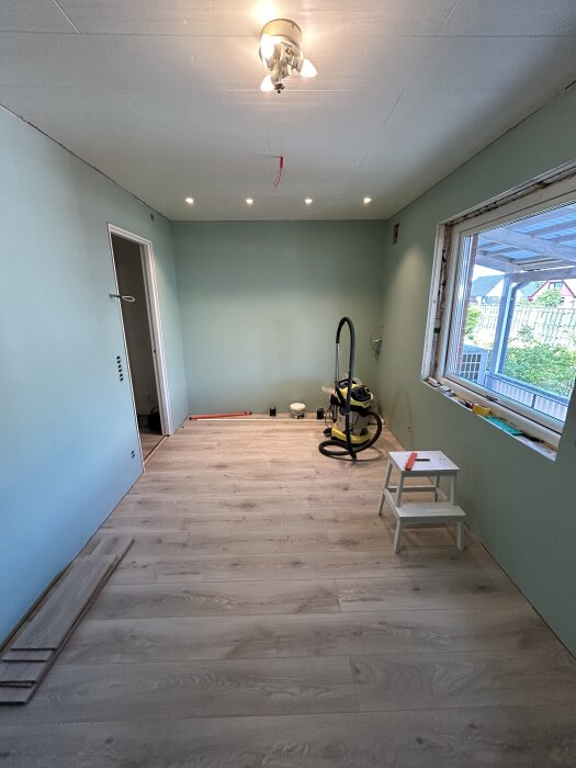 Rum med nylagt ljust trägolv, grönmålade väggar, taklampa och spotlights. En stege och dammsugare syns. Utsikt genom fönster.