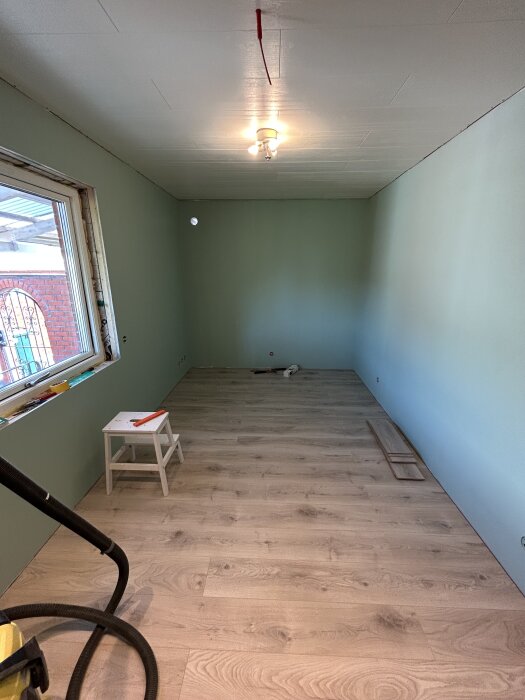 Nyinstallerat trägolv i ett rum med ljusgrönmålade väggar och enkel taklampa. Litet vitt stegverktyg och verktyg på golvet.