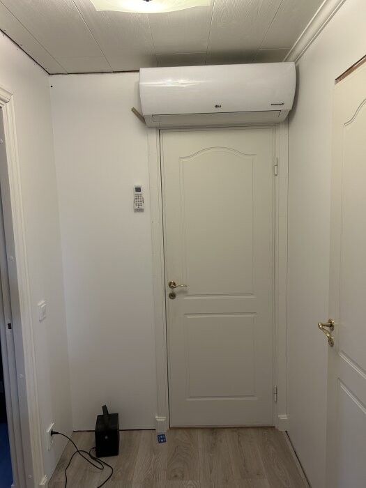 Luftvärmepump installerad ovanför en vit dörr i ett rum med parkettgolv. En fjärrkontroll sitter på väggen bredvid dörren.
