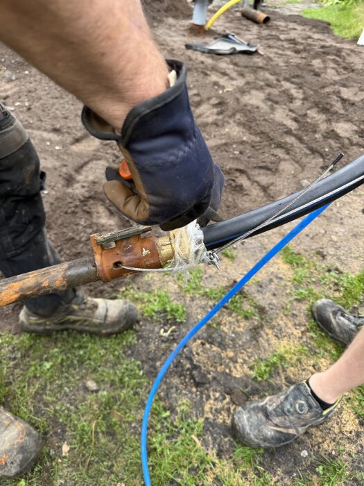 En person med handskar hanterar en gammal adapter med pågängat lyftrör utomhus, där det syns mark med verktyg och gräs.