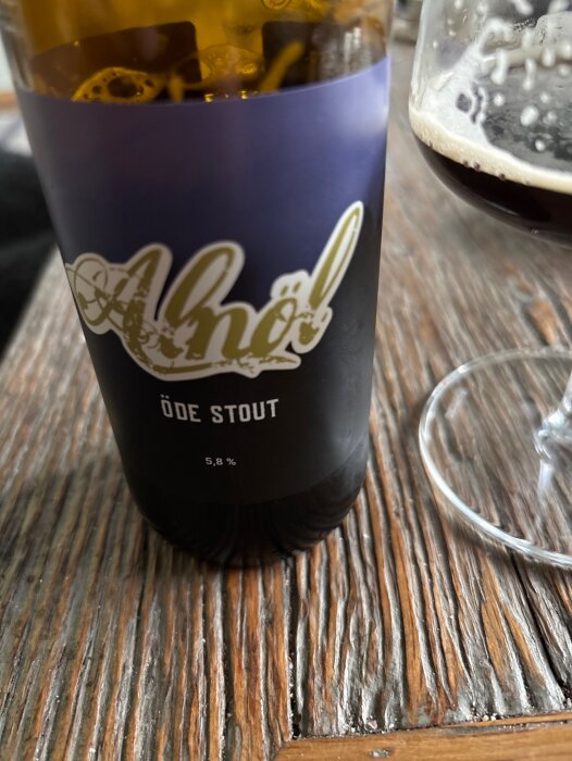Flaska med etikett "Alnöl Öde Stout 5,8%" på ett slitet träbord, bredvid ett halvfullt ölglas.