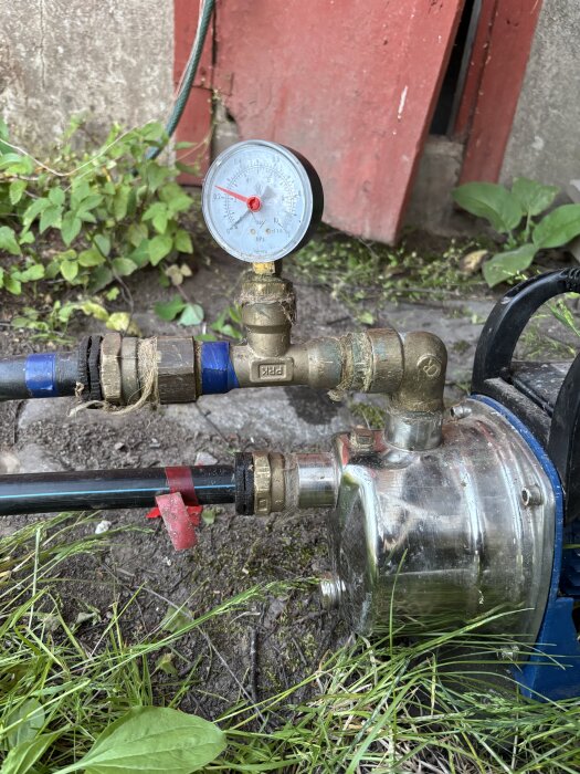 Vattenpump med rör och tryckmätare i trädgårdsmiljö, slangar svåra att demontera.