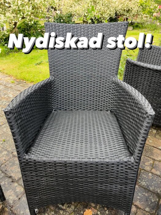 En nytvättad utomhusstol av flätat material står på en stenlagd uteplats med grönska i bakgrunden. Texten "Nydiskad stol!" finns på bilden.
